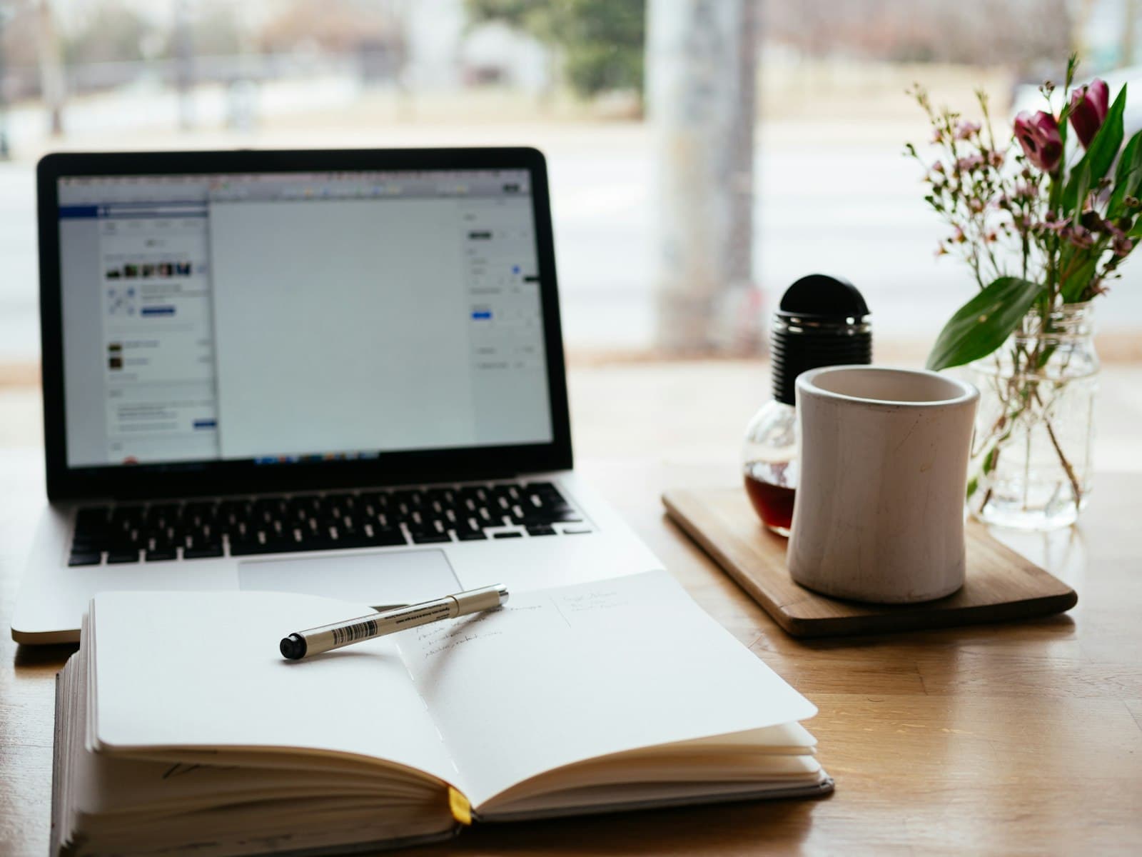 Tidy workspace with a laptop, open notebook, pen, mug and flowers by a window